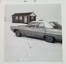 Old black and white photo of a winter scene, with a car from the 1960s and a small cottage in the background.